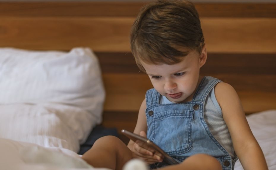 Kind sitzt im Bett und schaut in Smartphone. 